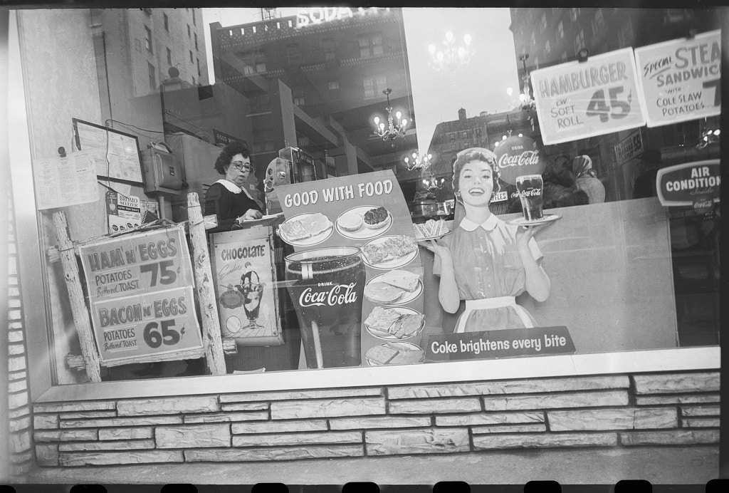 Woman at cash register viewed through diner show window with buildings reflected, 1959