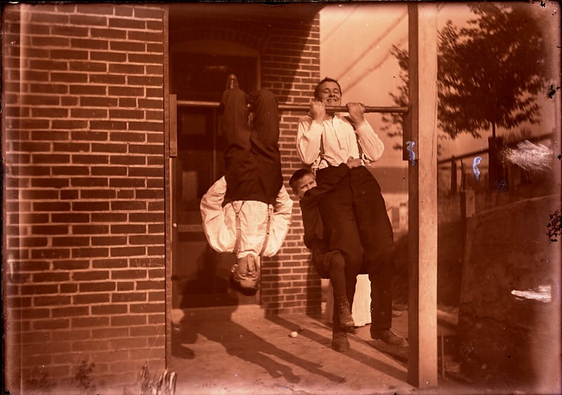 Bill Luetke and Otto Kroeger do gymnastics on a pull-up bar on a porch. One hangs upside down and the other has a small boy hanging on to him and he does a chin-up.