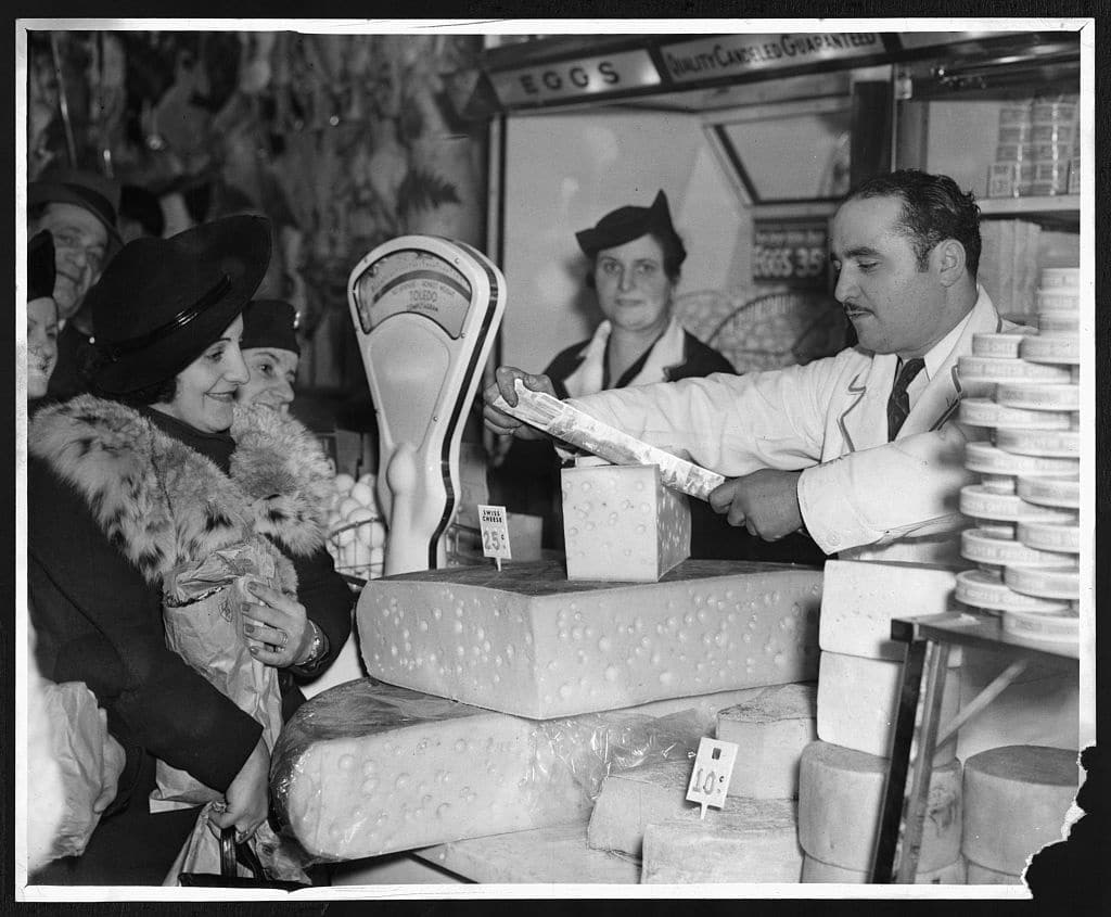 Vendor at the First Avenue Market slicing cheese for a customer (1938- New York)