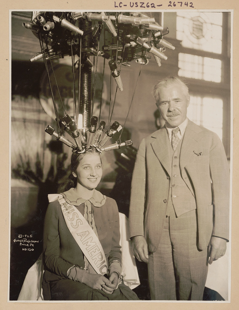 Miss America receives a permanent wave- Norma Smallwood, Miss America 1926, seated, with hair attached to electric permanent wave equipment, with man standing beside her. (1926 Oct. 14)