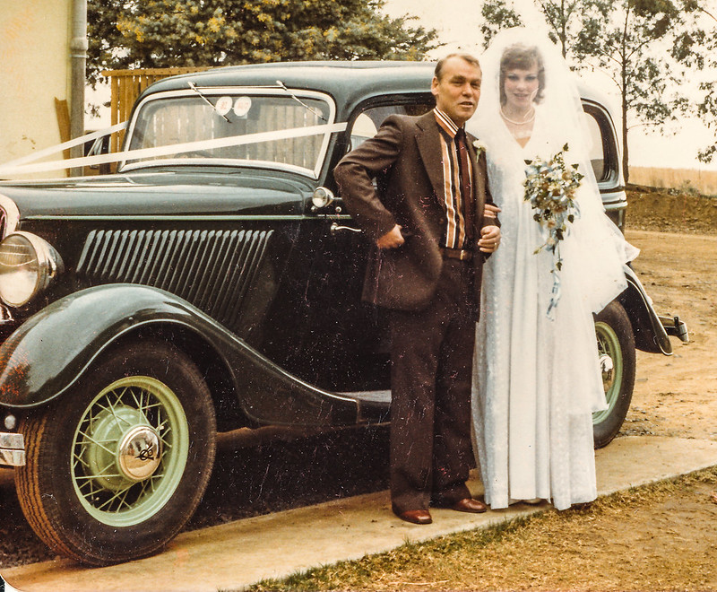 a man in a suit posing beside a bride. Behind them is a vintage car. With her bouquet still in her hand, they’re still on the way to her wedding.