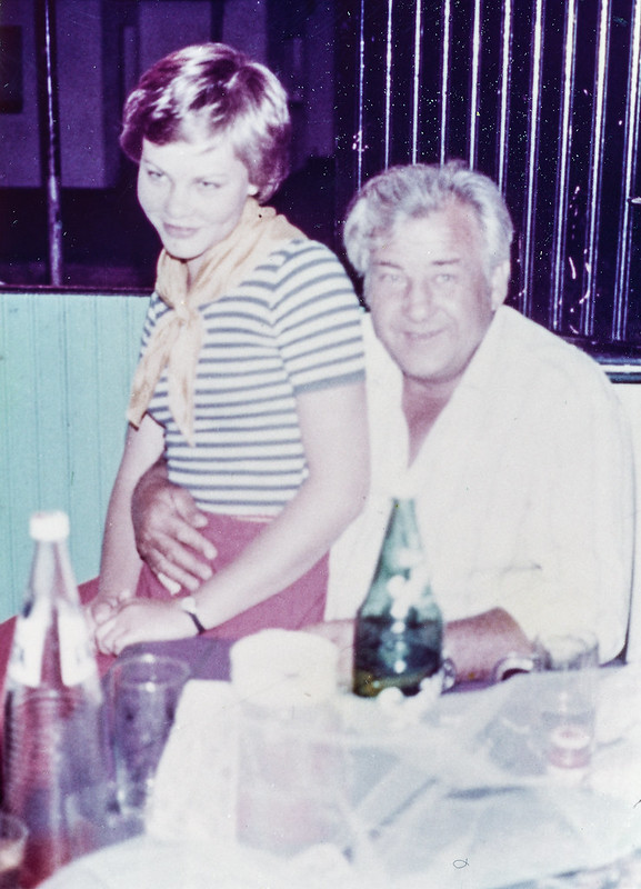 A younger woman sitting on the lap of an old man. The alcohol and bottles in front of them also show that they had been drinking