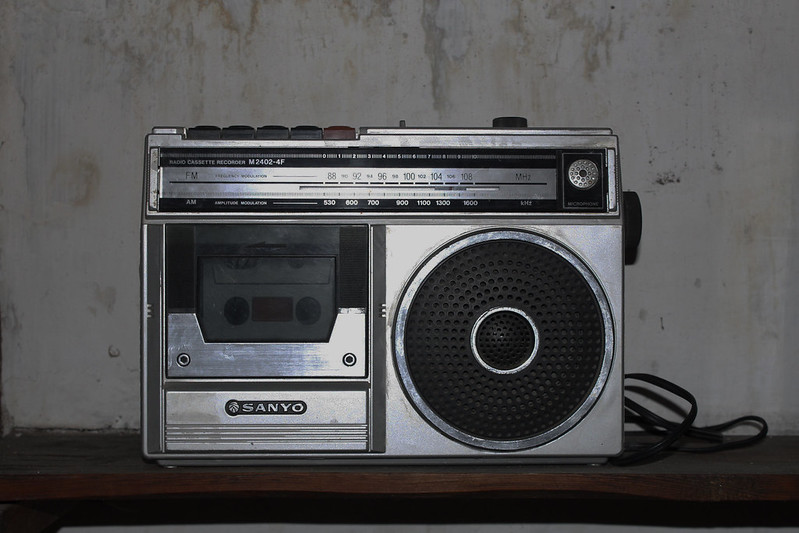 a 1970s radio cassette displayed in US living rooms. Brand is Sanyo. A radio cassette is a device that combines a radio receiver (AM/FM) with a cassette tape player/recorder. It allows users to listen to radio broadcasts and also play and record audio from cassette tapes. These devices were popular in the past and are sometimes referred to as boomboxes or portable cassette players with radio.