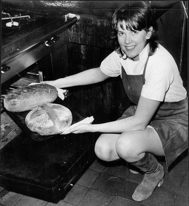 This image from the late Sixties or early Seventies shows celebrity chef Prue Leith at work in her kitchen. She is wearing a dark-coloured apron over a short, white work dress and - perhaps incongruously - knee-length boots. She is removing two freshly-baked loaves of bread from an oven.